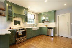 A kitchen with green cabinets, a stainless steel gas stove, and dishwasher, white countertops, and light wood flooring. There is a window above the sink and modern lighting fixtures.