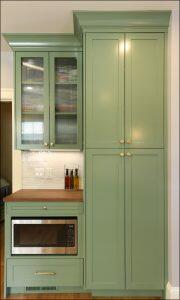 A kitchen with tall green cabinets, a built-in microwave oven, a wood countertop, and a white tile backsplash with bottles of cooking oil.