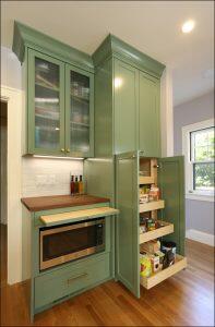 A kitchen with green cabinetry. The cabinets include a glass-front upper unit, a built-in microwave, and a pull-out pantry containing various food items. A window on the right lets in natural light.