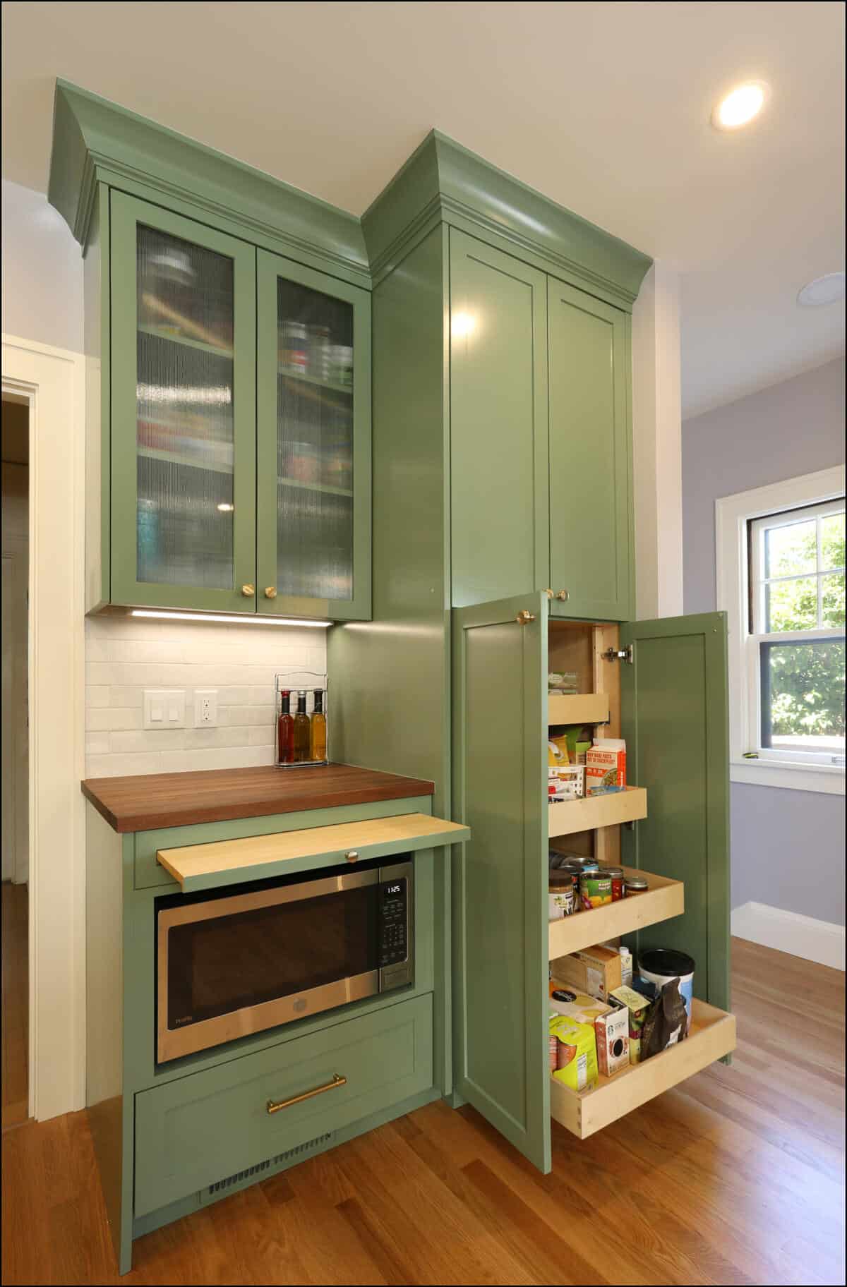 A kitchen with green cabinetry. The cabinets include a glass-front upper unit, a built-in microwave, and a pull-out pantry containing various food items. A window on the right lets in natural light. A kitchen with green cabinetry. The cabinets include a glass-front upper unit, a built-in microwave, and a pull-out pantry containing various food items. A window on the right lets in natural light.
