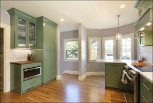 A modern kitchen with green cabinets, wooden flooring, a built-in microwave, and a countertop with a hanging pendant light. There are large windows providing natural light.