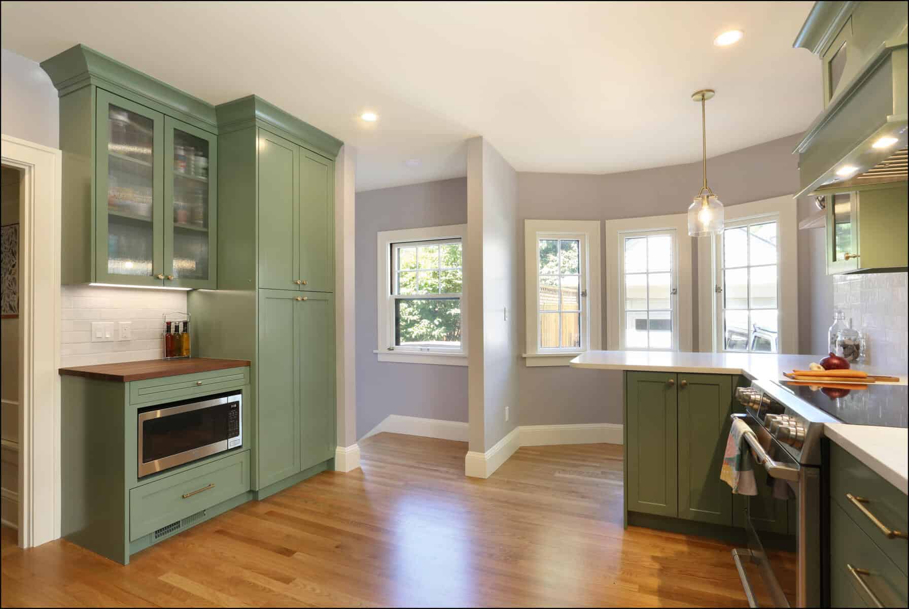 A modern kitchen with green cabinets, wooden flooring, a built-in microwave, and a countertop with a hanging pendant light. There are large windows providing natural light. A modern kitchen with green cabinets, wooden flooring, a built-in microwave, and a countertop with a hanging pendant light. There are large windows providing natural light.
