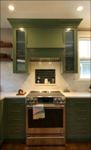 Modern kitchen with green cabinetry, stainless steel oven, and black backsplash. White subway tile surrounds the cooking area, with wooden shelves on the left and a window above the countertop on the right.