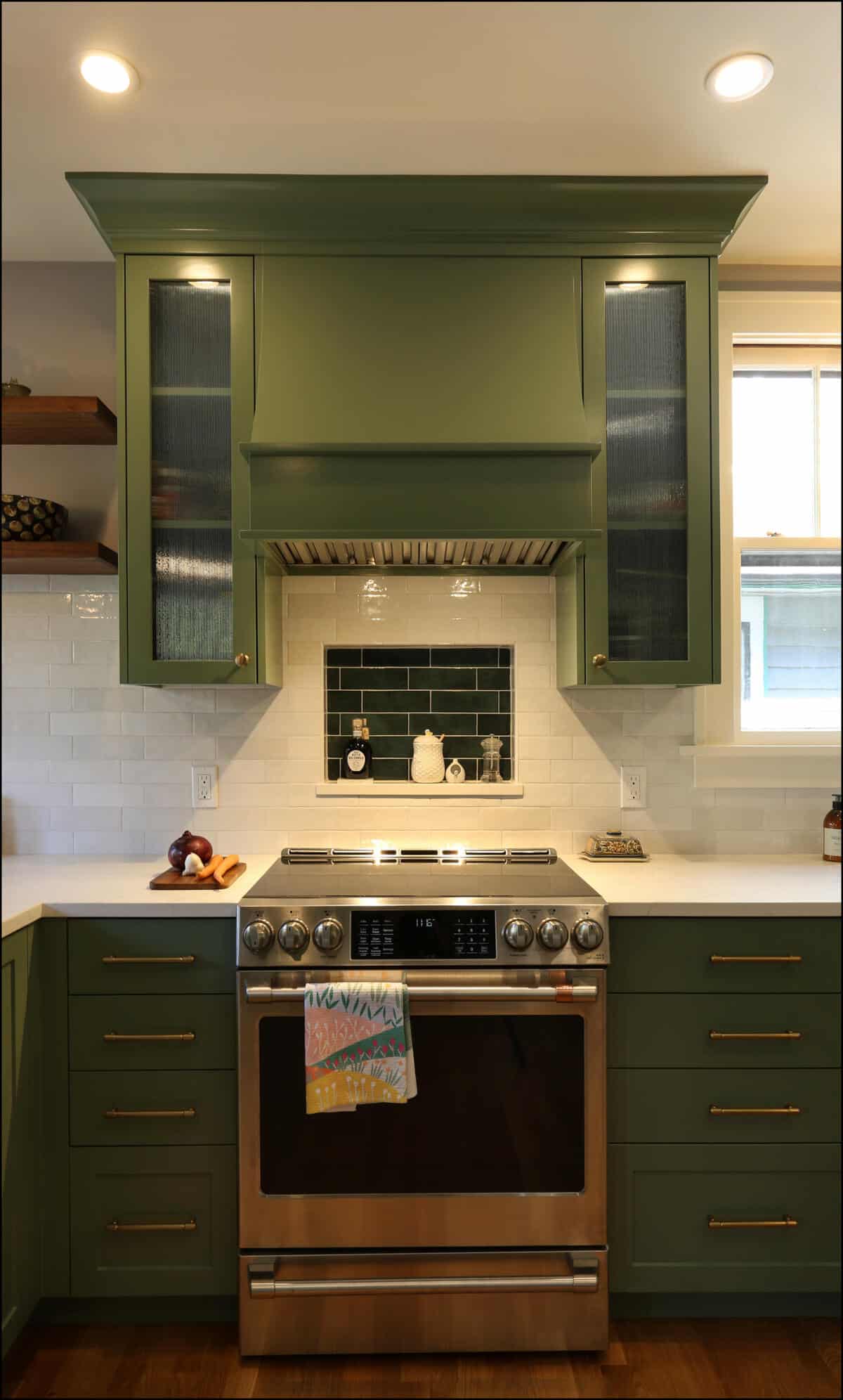 Modern kitchen with green cabinetry, stainless steel oven, and black backsplash. White subway tile surrounds the cooking area, with wooden shelves on the left and a window above the countertop on the right. Modern kitchen with green cabinetry, stainless steel oven, and black backsplash. White subway tile surrounds the cooking area, with wooden shelves on the left and a window above the countertop on the right.