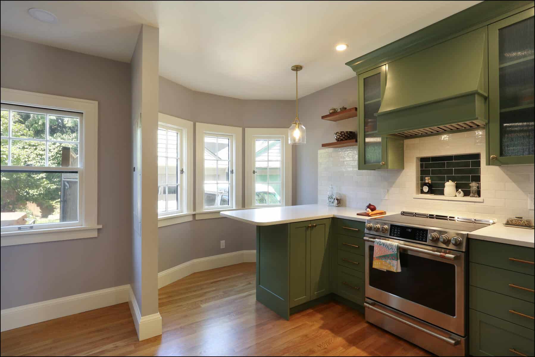 A modern kitchen with green cabinets, stainless steel appliances, a white countertop, a pendant light, and a large bay window. A modern kitchen with green cabinets, stainless steel appliances, a white countertop, a pendant light, and a large bay window.