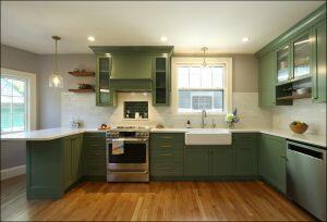 A modern kitchen with green cabinets, white countertops, stainless steel appliances, and a farm-style sink. A pendant light hangs over the island. The floor is hardwood.