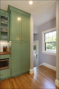 A kitchen corner featuring green cabinets, a microwave, and a door with a window to the outside. The room has wooden floors and a small window next to the door.