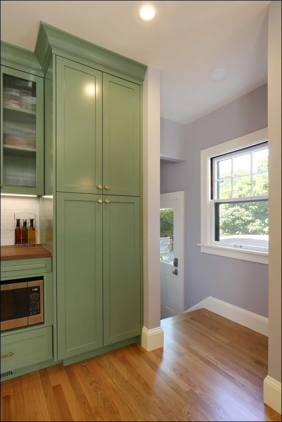A kitchen corner featuring green cabinets, a microwave, and a door with a window to the outside. The room has wooden floors and a small window next to the door. A kitchen corner featuring green cabinets, a microwave, and a door with a window to the outside. The room has wooden floors and a small window next to the door.