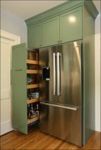 A modern kitchen with green cabinetry, a double-door stainless steel refrigerator, and an open pantry with wooden pull-out shelves stocked with various food items.