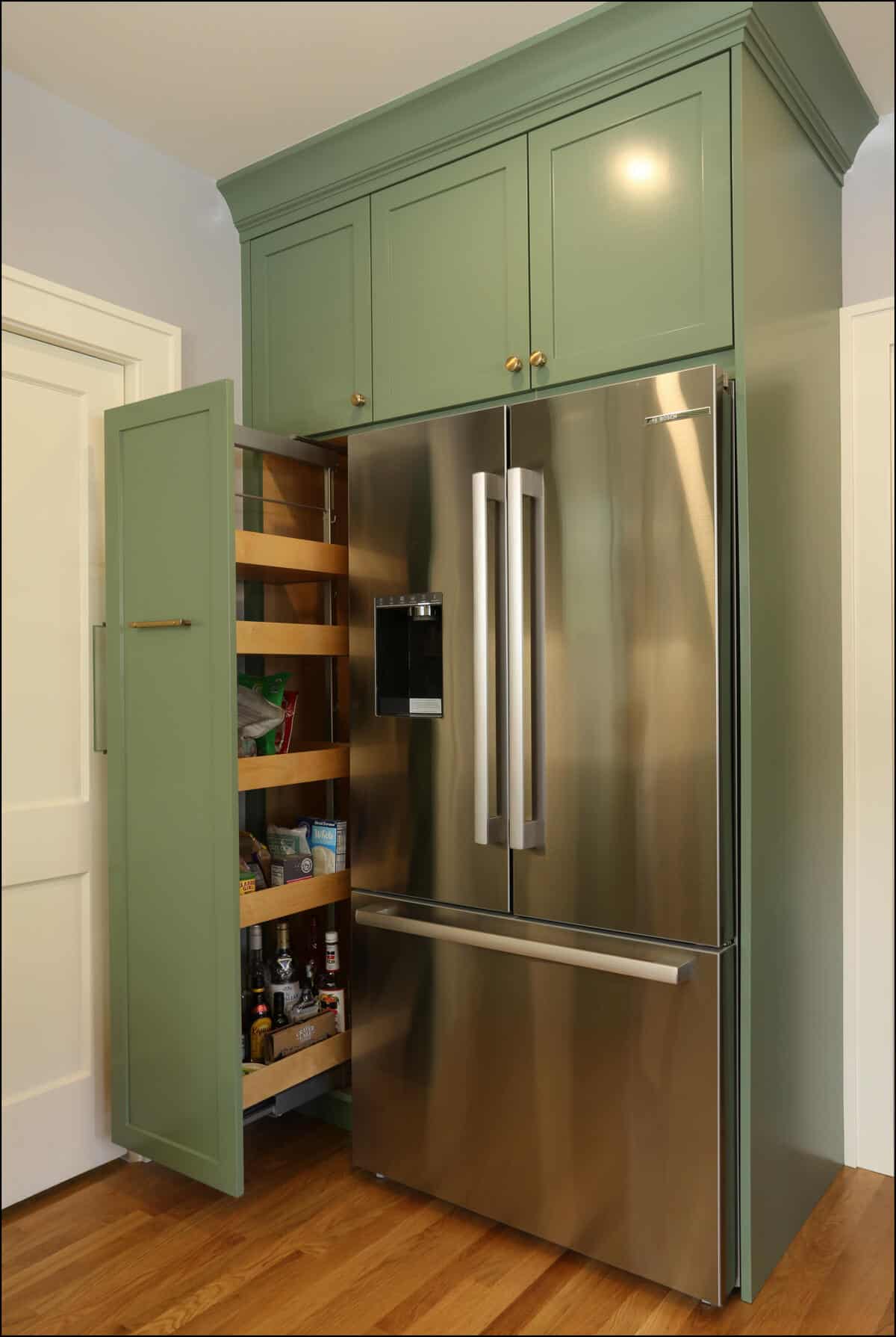 A modern kitchen with green cabinetry, a double-door stainless steel refrigerator, and an open pantry with wooden pull-out shelves stocked with various food items. A modern kitchen with green cabinetry, a double-door stainless steel refrigerator, and an open pantry with wooden pull-out shelves stocked with various food items.