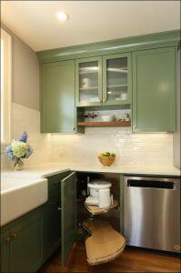 A kitchen with green cabinets, white countertops, a dishwasher, and a sink. The lower cabinet door is open, revealing a pull-out shelf with a food processor. A bowl of apples and vases are on the counter.