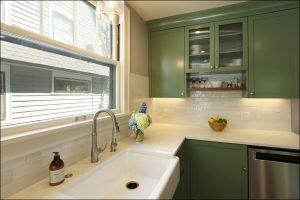 A kitchen with green cabinets, white countertops, a stainless steel sink, and a large window. A bowl of lemons and a vase of flowers are on the counter next to a soap dispenser.