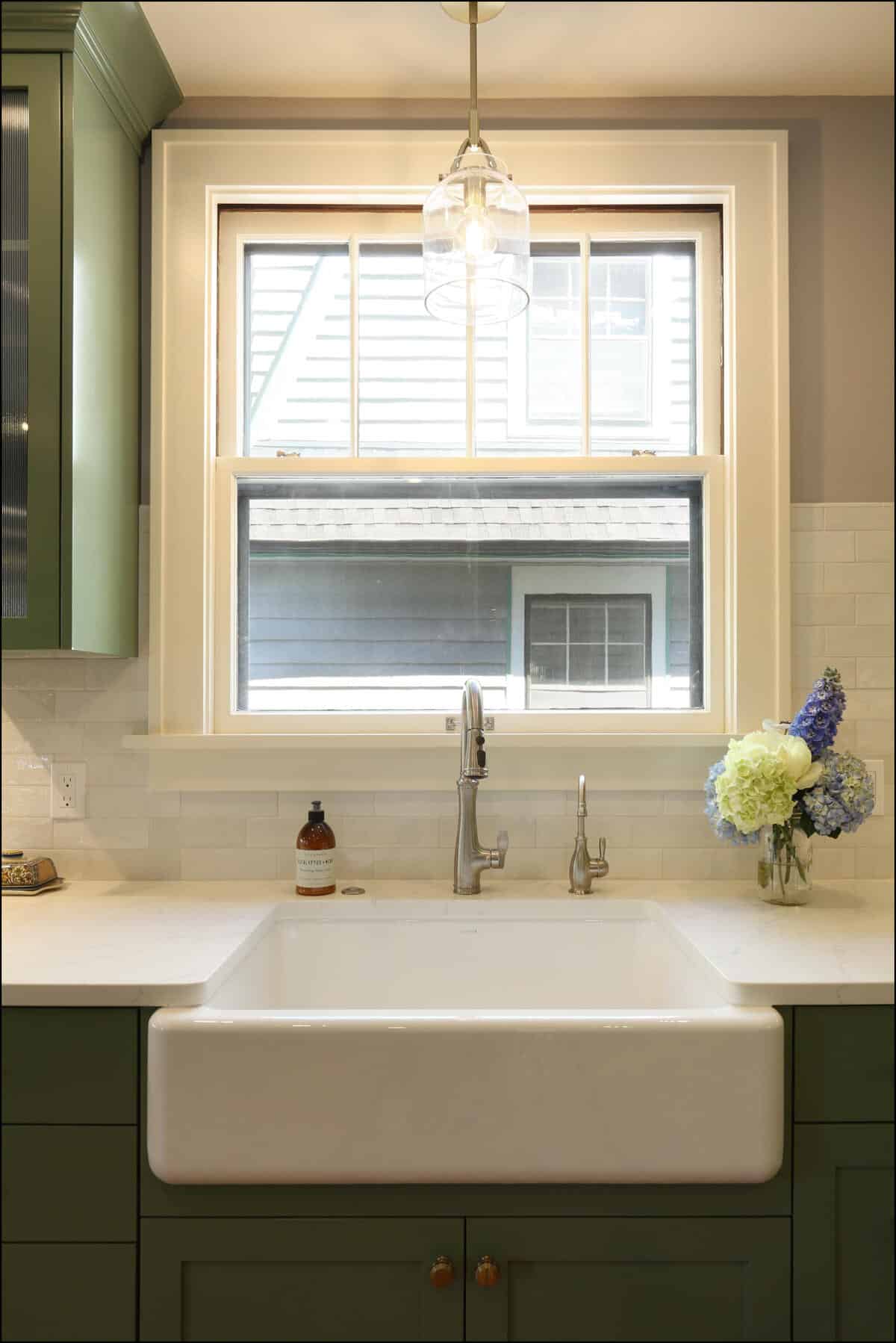 A kitchen sink under a double-hung window with a single pendant light above. There's a soap dispenser and a vase with flowers on the countertop next to the sink. A kitchen sink under a double-hung window with a single pendant light above. There's a soap dispenser and a vase with flowers on the countertop next to the sink.