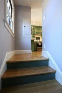 A short wooden staircase leads to a kitchen with green cabinets, a range hood, and a stove, seen from a hallway with a window and electrical panel on the wall.
