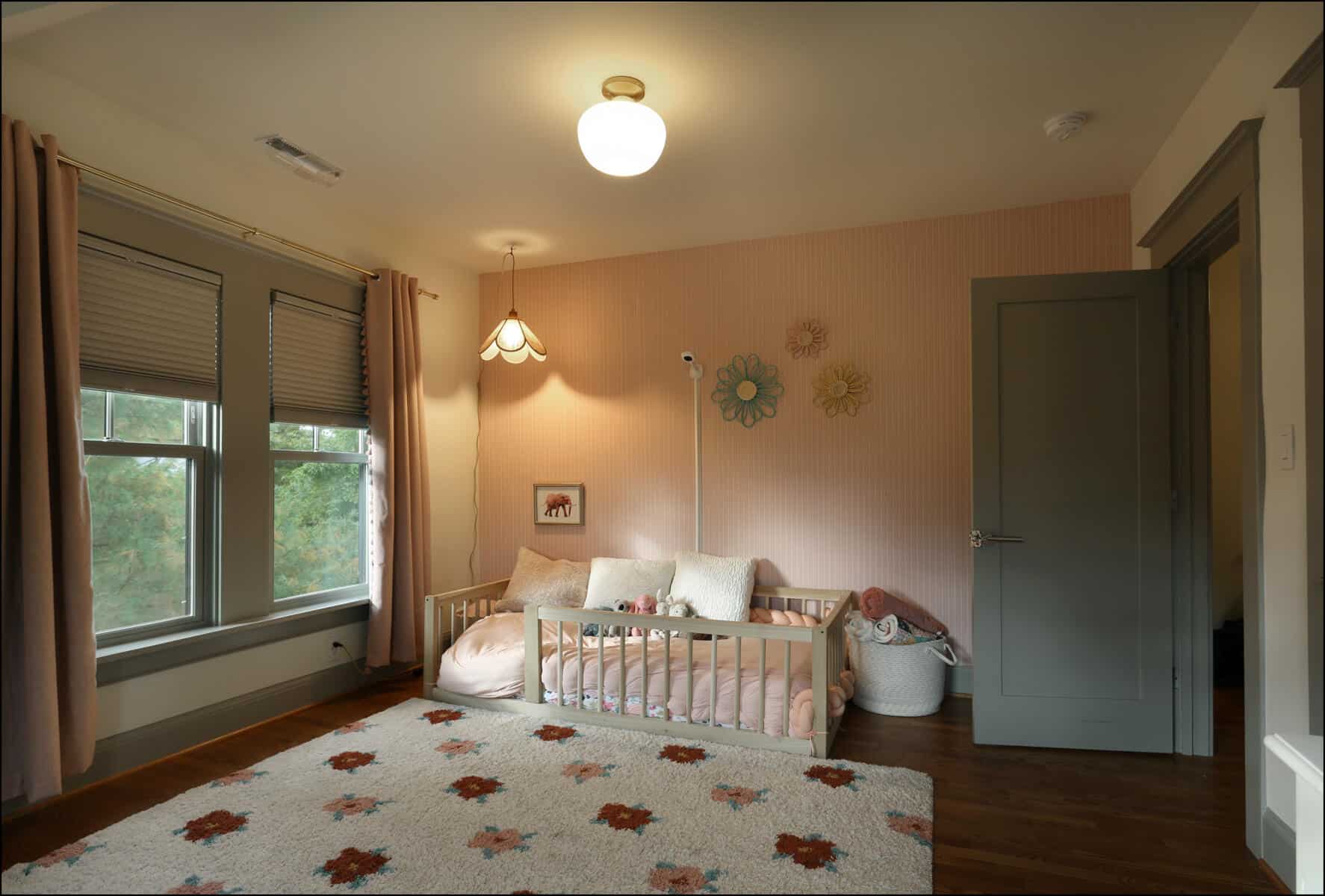 A cozy child's room with a bed, a basket of toys, a large rug, and floral wall decorations. Two large windows and a ceiling light illuminate the space. Brightly lit nursery featuring a crib, decorative wall elements, and large windows, showcasing a modern and cozy design ideal for whole house remodeling projects by ReCraft Home Remodeling in Portland.