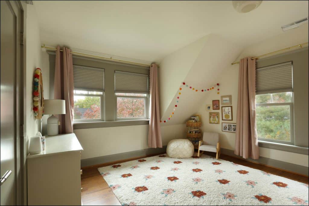 A cozy room with light pink curtains, colorful wall decorations, a white rug with red patterns, and framed pictures. A small chair and pouf are in the corner near two large windows. A cozy room with light pink curtains, colorful wall decorations, a white rug with red patterns, and framed pictures. A small chair and pouf are in the corner near two large windows.