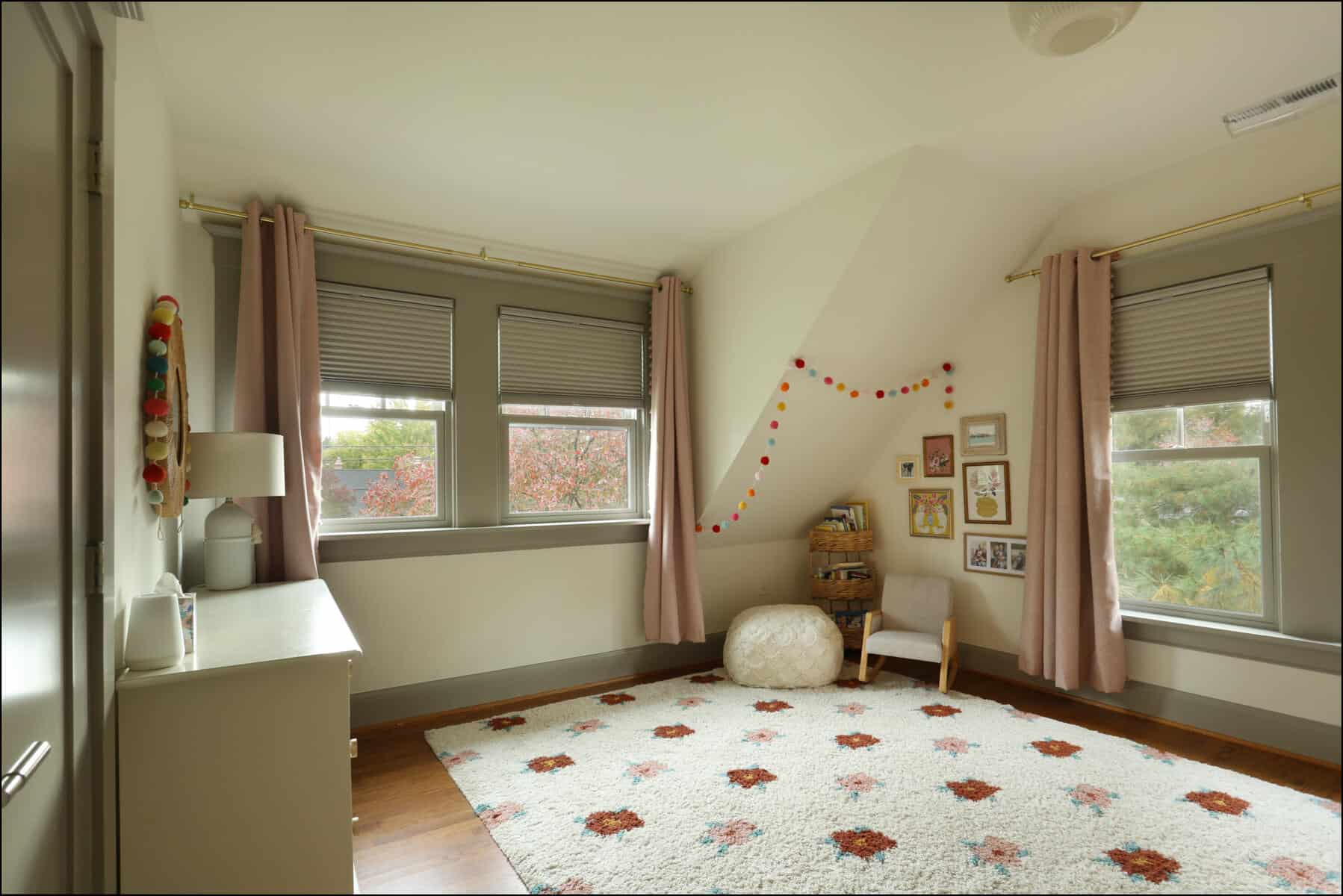 A cozy room with light pink curtains, colorful wall decorations, a white rug with red patterns, and framed pictures. A small chair and pouf are in the corner near two large windows. Cozy children's room featuring a plush rug, light curtains, and a mix of decorative elements, showcasing ReCraft Home Remodeling's interior design expertise in whole house remodels in Portland, Oregon.