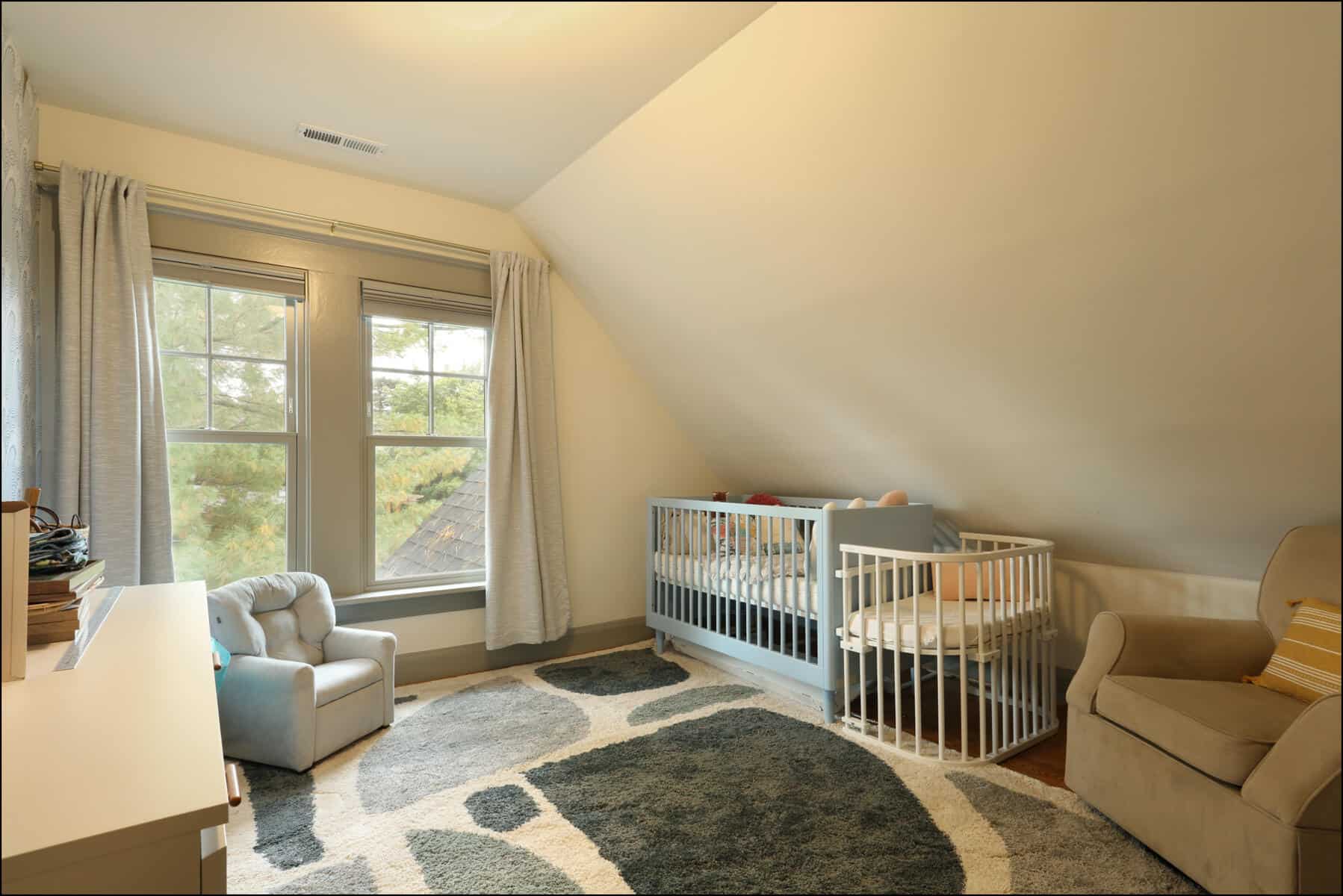 A cozy nursery with a crib, armchair, and plush chair. A rug covers part of the floor. There are two windows with curtains on the left wall. Cozy nursery room featuring a crib, comfortable armchair, and large windows with natural light, showcasing a whole house remodel by ReCraft Home Remodeling in Portland, Oregon.