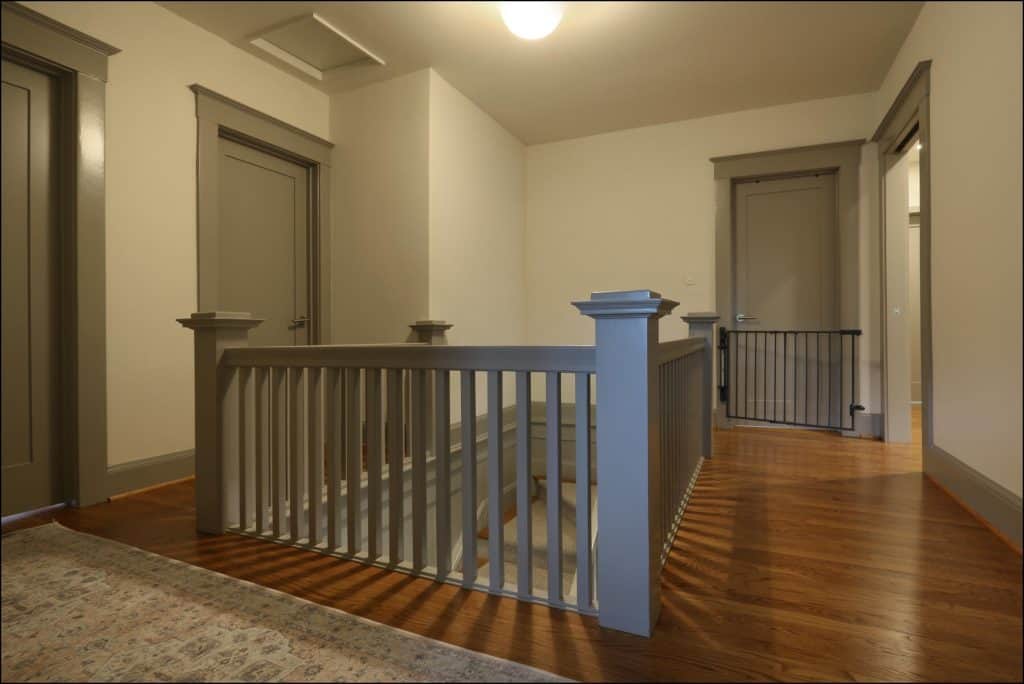 A hallway with wooden floors, two closed doors, and a staircase behind a railing. Walls and door frames are painted in neutral colors. A hallway with wooden floors, two closed doors, and a staircase behind a railing. Walls and door frames are painted in neutral colors.