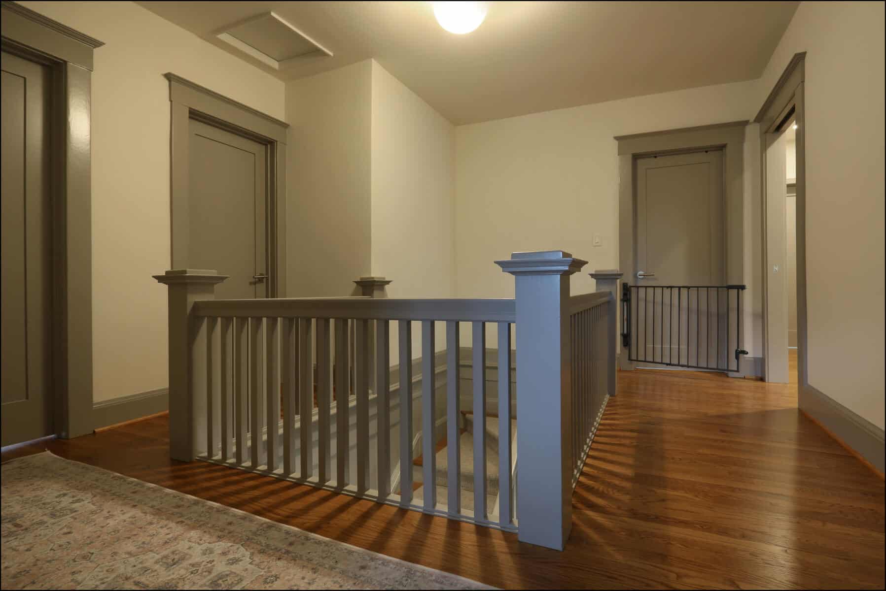 A hallway with wooden floors, two closed doors, and a staircase behind a railing. Walls and door frames are painted in neutral colors. Interior view of a remodeled hallway featuring a wooden railing, hardwood flooring, and multiple doorways, showcasing ReCraft Home Remodeling's design expertise in whole house renovations in Portland.