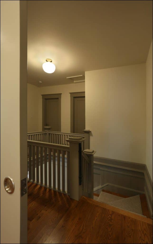A dimly lit hallway with two closed doors and a wooden staircase with a railing, viewed from an open doorway. A dimly lit hallway with two closed doors and a wooden staircase with a railing, viewed from an open doorway.