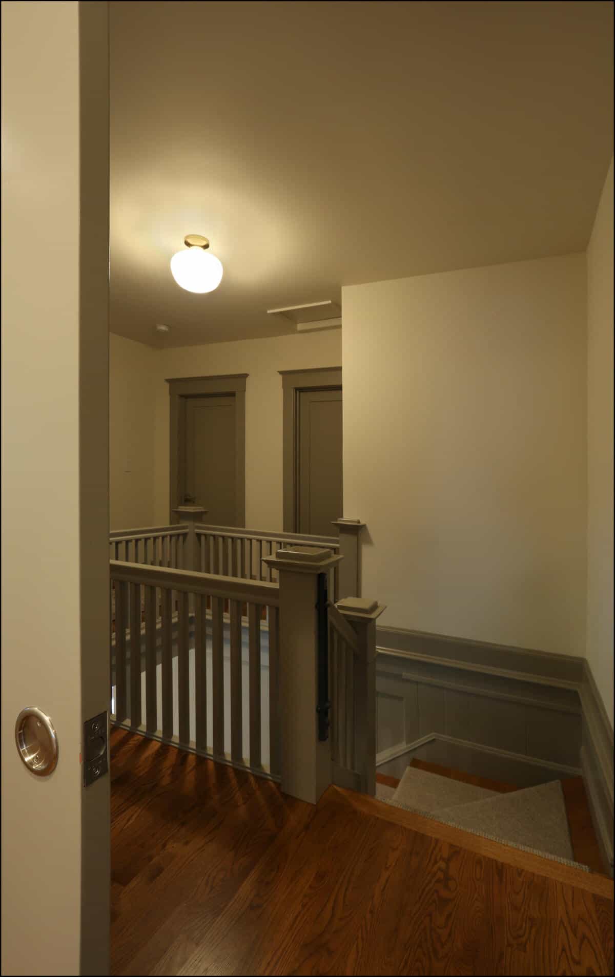 A dimly lit hallway with two closed doors and a wooden staircase with a railing, viewed from an open doorway. Interior view of a well-lit staircase landing featuring wooden flooring, gray railings, and two closed doors, showcasing the design elements of a whole house remodel by ReCraft Home Remodeling in Portland, OR.