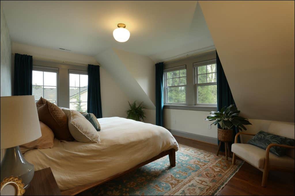 A cozy bedroom with a bed, pillows, an armchair, and plants. The room has slanted ceilings, natural light from windows, and a patterned rug on a wooden floor. A cozy bedroom with a bed, pillows, an armchair, and plants. The room has slanted ceilings, natural light from windows, and a patterned rug on a wooden floor.