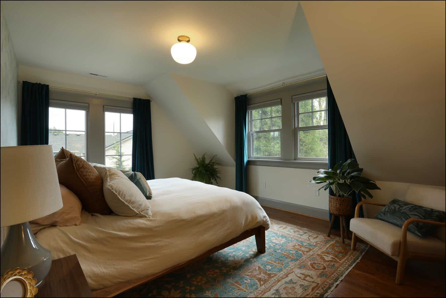 A cozy bedroom with a bed, pillows, an armchair, and plants. The room has slanted ceilings, natural light from windows, and a patterned rug on a wooden floor. Cozy bedroom featuring a bed with decorative pillows, a stylish lamp, large windows with curtains, and a comfortable chair, showcasing a modern whole house remodel by ReCraft Home Remodeling in Portland, Oregon.