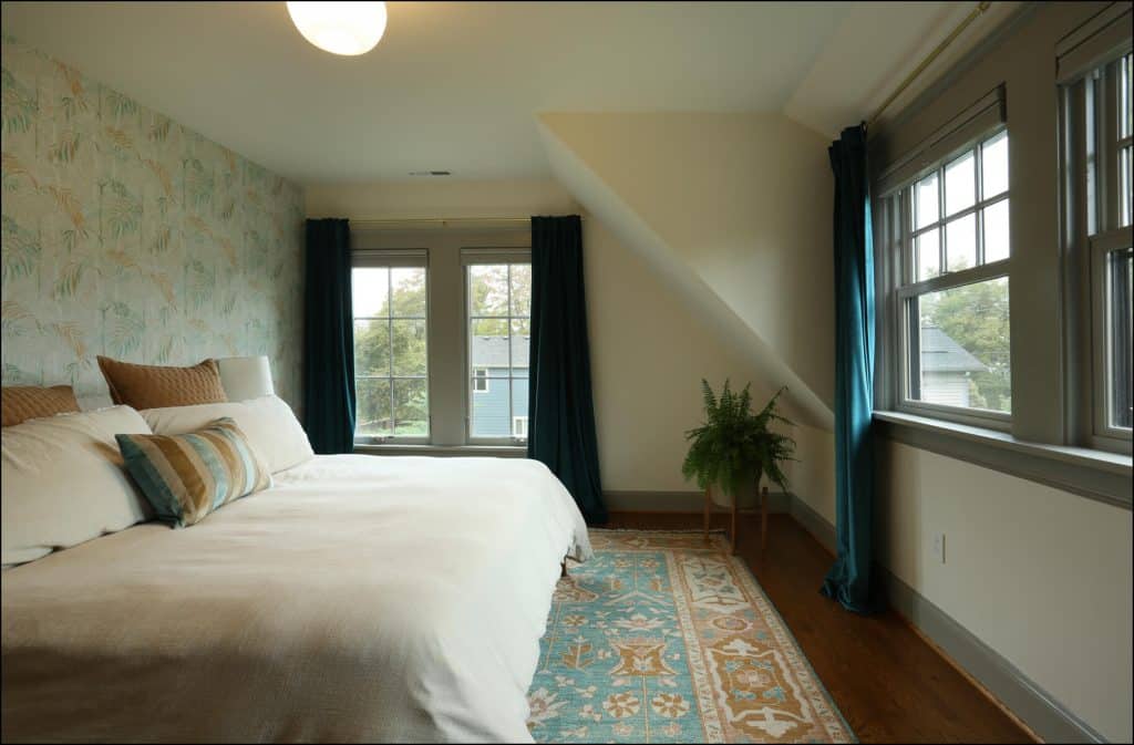 A bedroom with a double bed, decorative pillows, a patterned rug, and a potted plant by the window. The room features floral wallpaper and dark curtains. A bedroom with a double bed, decorative pillows, a patterned rug, and a potted plant by the window. The room features floral wallpaper and dark curtains.