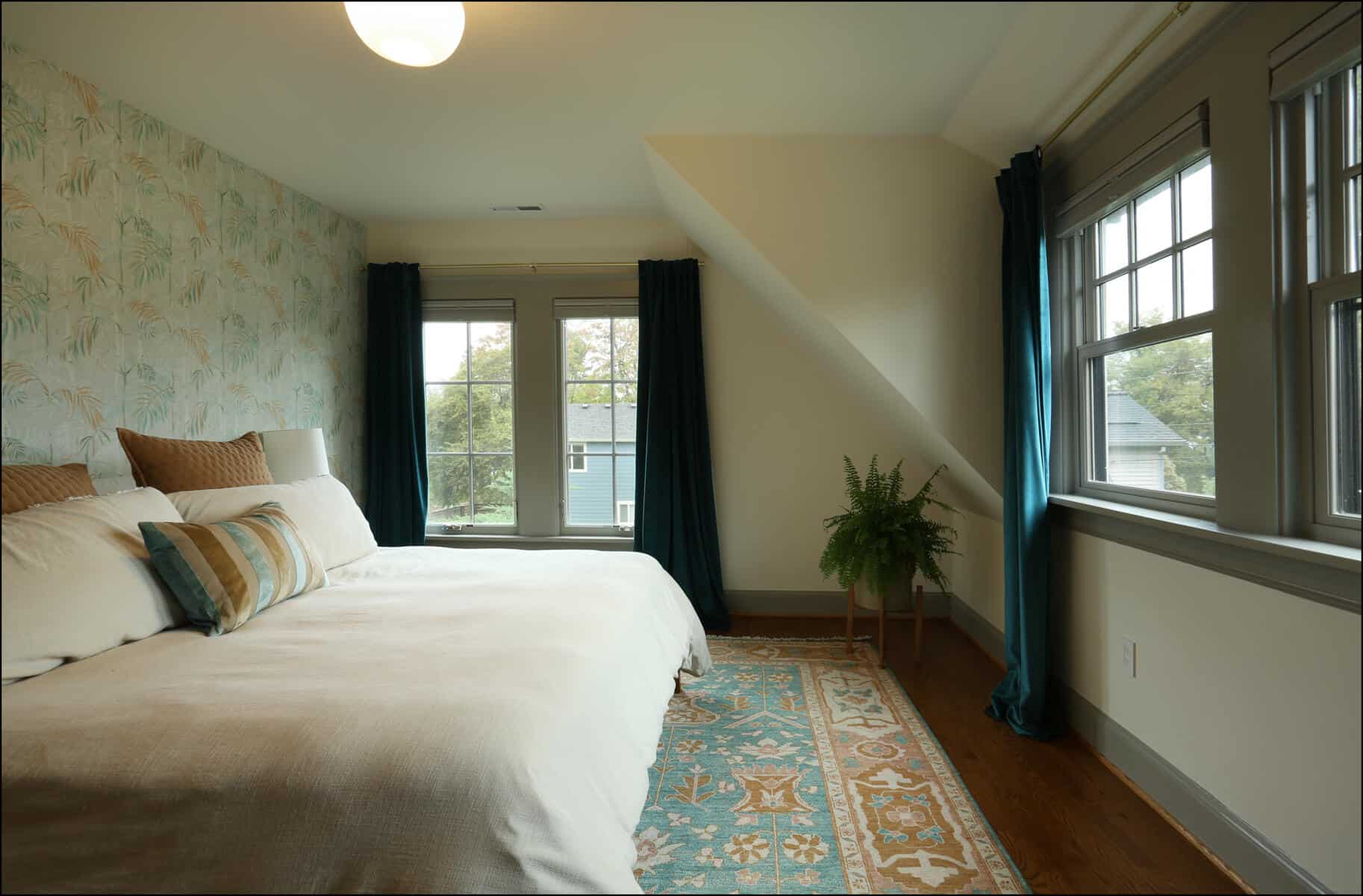 A bedroom with a double bed, decorative pillows, a patterned rug, and a potted plant by the window. The room features floral wallpaper and dark curtains. Cozy bedroom featuring a large bed with decorative pillows, elegant wallpaper, and natural light from windows, showcasing ReCraft Home Remodeling's interior design capabilities in whole house remodels.