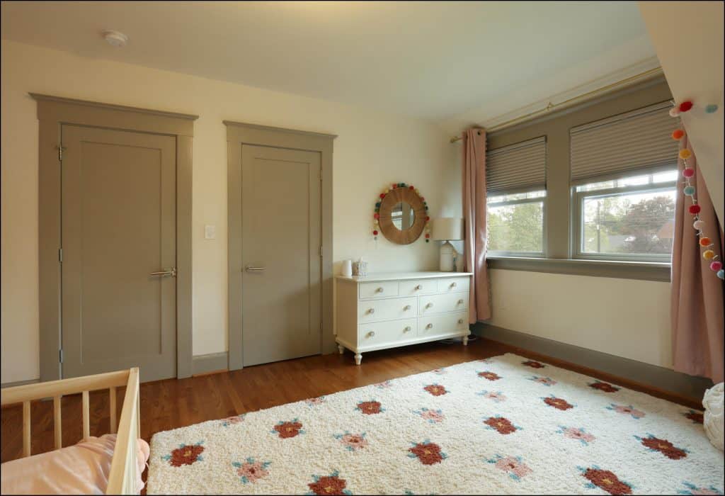 A cozy children's bedroom with a small bed, dresser, and mirror. The room has beige walls, wooden flooring, and a rug with floral patterns. Two closed doors and a window with curtains are visible. A cozy children's bedroom with a small bed, dresser, and mirror. The room has beige walls, wooden flooring, and a rug with floral patterns. Two closed doors and a window with curtains are visible.