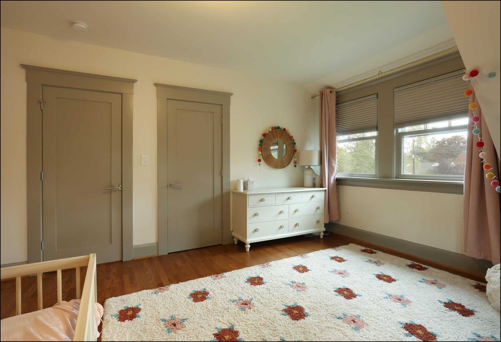 A cozy children's bedroom with a small bed, dresser, and mirror. The room has beige walls, wooden flooring, and a rug with floral patterns. Two closed doors and a window with curtains are visible. Cozy nursery featuring a crib, floral-patterned rug, dresser, and large windows with soft curtains, showcasing a warm and inviting design for a whole house remodel in Portland.