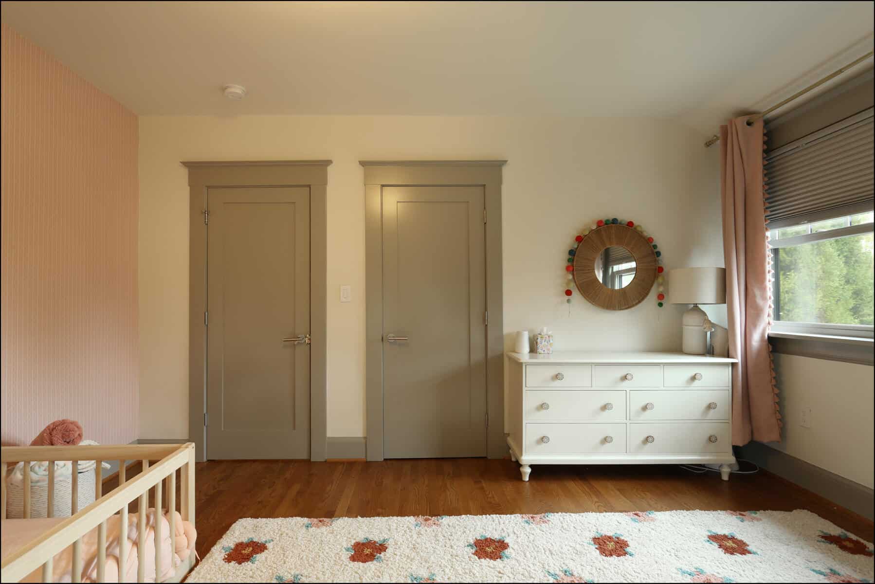 A nursery room features a crib, dresser, mirror, and two closed doors. A rug with floral patterns lies on the wooden floor, and a window with blinds and curtains lets in natural light. Brightly lit nursery featuring a crib, white dresser, and mirror, with pink accent wall and wooden flooring, showcasing ReCraft Home Remodeling's interior design work in whole house remodeling projects.