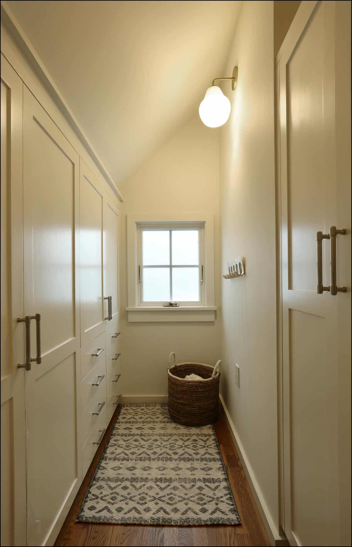 A narrow, well-lit hallway with built-in cabinets on the left, a small window at the end, a patterned rug on the wooden floor, and a wicker basket in the corner. Narrow hallway featuring built-in cabinetry, a window allowing natural light, a patterned area rug, and a decorative basket, showcasing a modern whole house remodel by ReCraft Home Remodeling in Portland, Oregon.