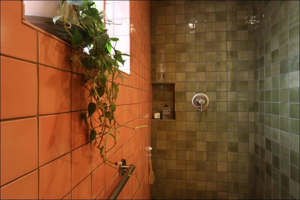 Shower area with green and orange tiles, featuring a wall-mounted showerhead, a small niche with toiletries, and a window with trailing plants. Shower area with green and orange tiles, featuring a wall-mounted showerhead, a small niche with toiletries, and a window with trailing plants.