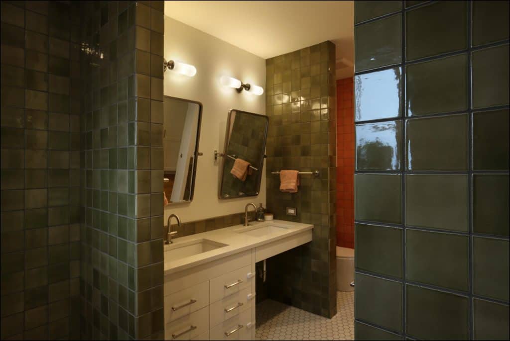 Bathroom with green tiled walls, double sinks, and illuminated mirrors. White cabinet below sinks and orange tiled section visible in the background. Bathroom with green tiled walls, double sinks, and illuminated mirrors. White cabinet below sinks and orange tiled section visible in the background.