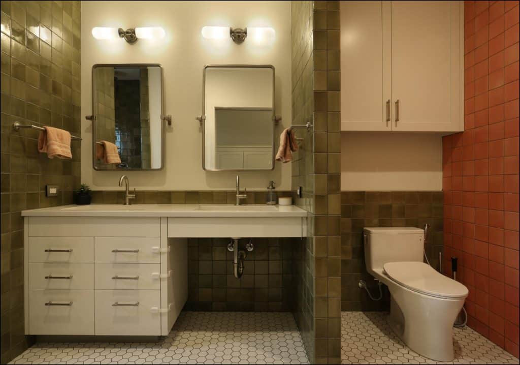 A modern bathroom with a dual sink vanity, two mirrors, a toilet, and green tiled walls. Towels hang on both sides of the vanity. Hexagonal floor tiles are white. A modern bathroom with a dual sink vanity, two mirrors, a toilet, and green tiled walls. Towels hang on both sides of the vanity. Hexagonal floor tiles are white.