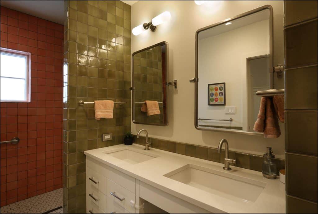 Bathroom with two sinks, large mirrors, and green tiled walls. Orange tiles and a window are visible in the adjacent shower area. Towels are hanging on the rack. Bathroom with two sinks, large mirrors, and green tiled walls. Orange tiles and a window are visible in the adjacent shower area. Towels are hanging on the rack.