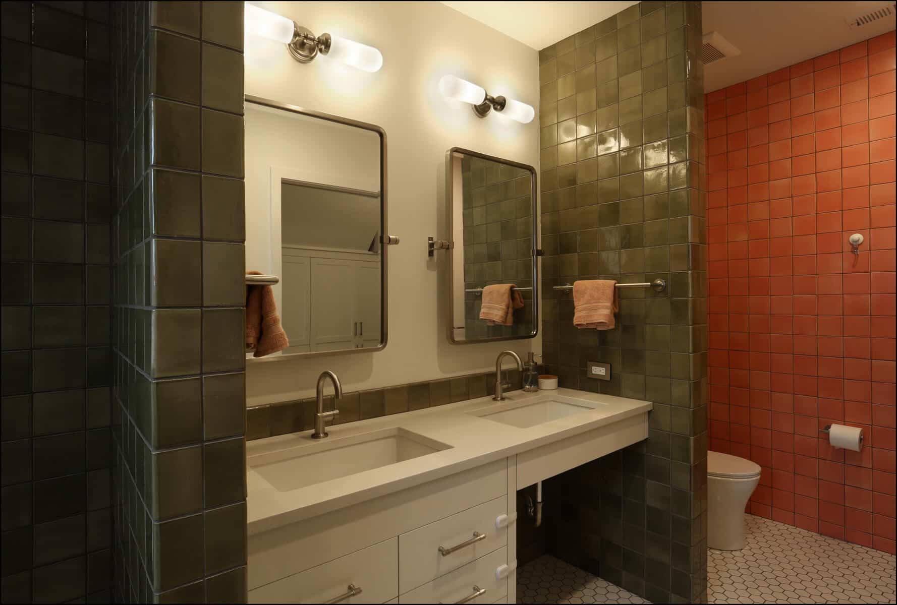 Modern bathroom featuring dual mirrors, green tiled walls, white vanity with sink, and orange accent wall.