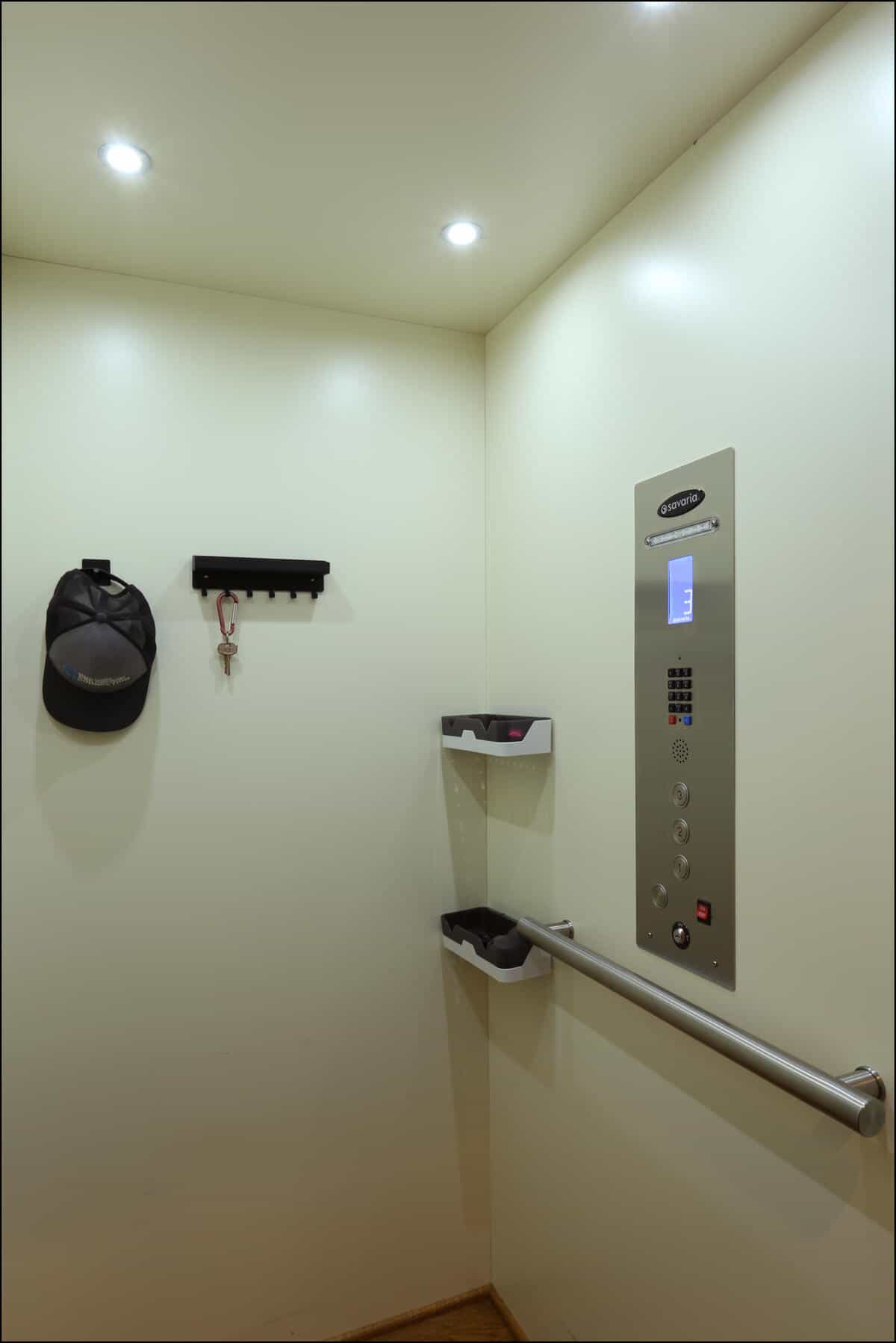 Interior of an elevator with a control panel, handrail, and two small shelves. A hat and keys hang on a wall hook. Elevator interior with control panel, wall-mounted shelves, and hat rack, showcasing modern design and functionality in a remodeled space.