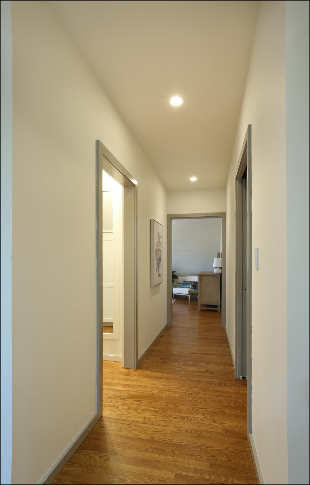 A hallway with wooden floors leads to a room with a bed and two lamps. Walls are cream-colored with a painting on the right. Modern hallway with hardwood flooring, featuring multiple doorways leading to rooms, illuminated by recessed lighting, showcasing the clean design typical of whole house remodeling by ReCraft Home Remodeling in Portland.