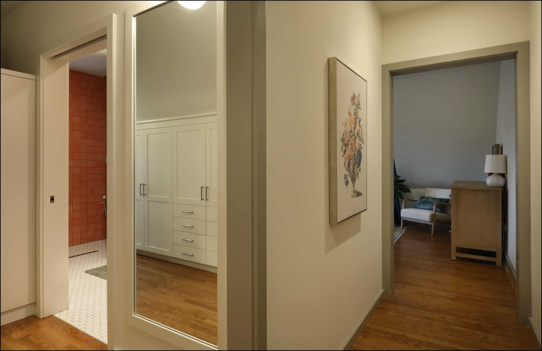 A hallway features a mirrored closet on the left, red-tiled bathroom, and a room with wooden flooring and furniture on the right. Hallway view showcasing a remodeled space with a large mirror, white cabinetry, and a glimpse of a cozy room in the background, emphasizing ReCraft Home Remodeling's design quality in whole house renovations.