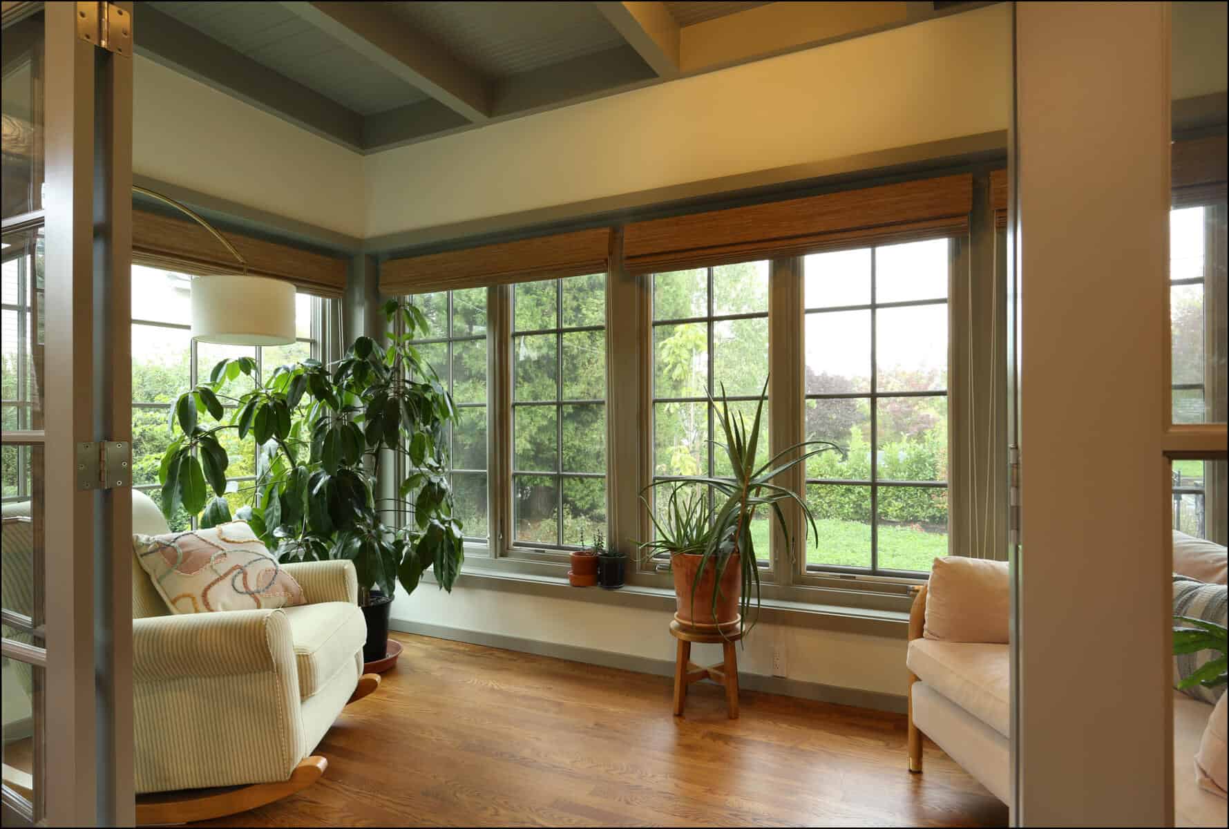 A cozy sunroom with large windows, green plants, a white armchair with a patterned pillow, and a beige chair on a wooden floor. Cozy sunlit living space featuring large windows, indoor plants, and comfortable seating, showcasing ReCraft Home Remodeling's whole house design in Portland, Oregon.