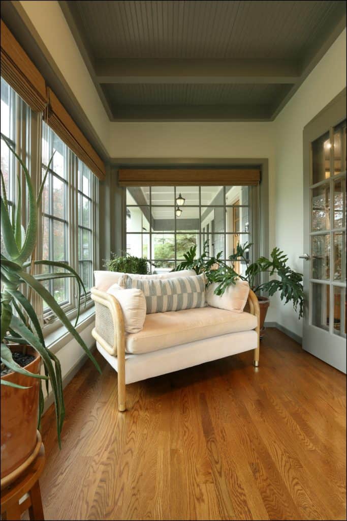 A cozy sunroom features a cushioned loveseat with striped pillows, surrounded by large potted plants, hardwood floor, and large windows with views of greenery. A cozy sunroom features a cushioned loveseat with striped pillows, surrounded by large potted plants, hardwood floor, and large windows with views of greenery.