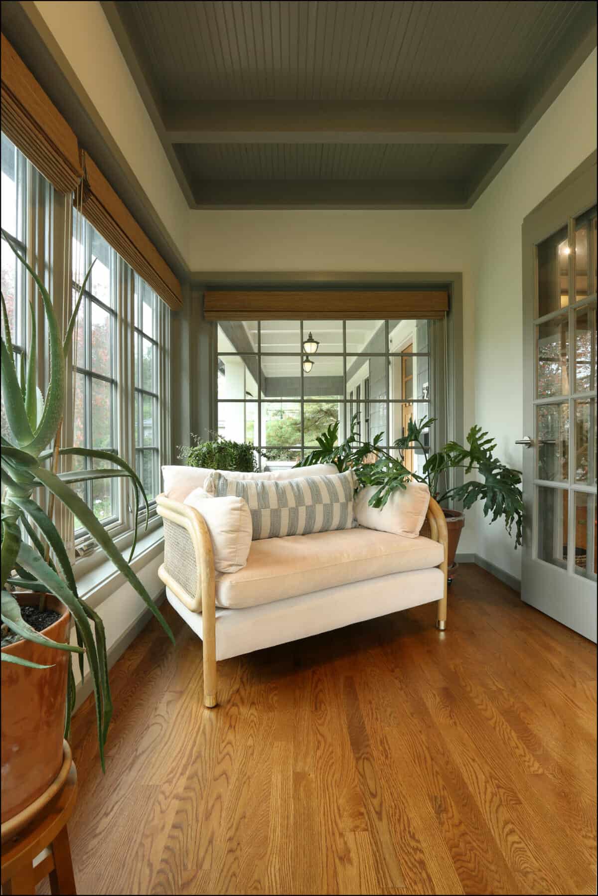 A cozy sunroom features a cushioned loveseat with striped pillows, surrounded by large potted plants, hardwood floor, and large windows with views of greenery. Cozy seating area featuring a light-colored sofa with decorative pillows, surrounded by large windows and indoor plants, showcasing a bright and inviting space in a whole house remodel by ReCraft Home Remodeling in Portland, Oregon.