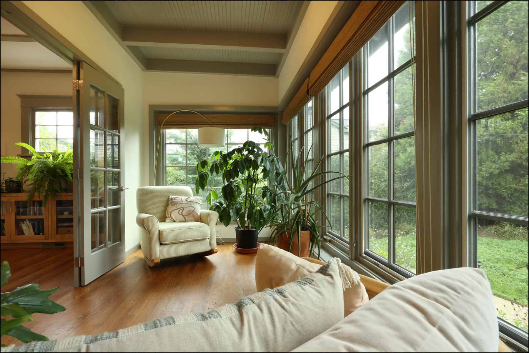 A cozy sunroom with large windows, an armchair, potted plants, and wooden floors overlooking a garden. Bright and inviting sunroom with large windows, comfortable seating, and indoor plants, showcasing a whole house remodel by ReCraft Home Remodeling in Portland, Oregon.