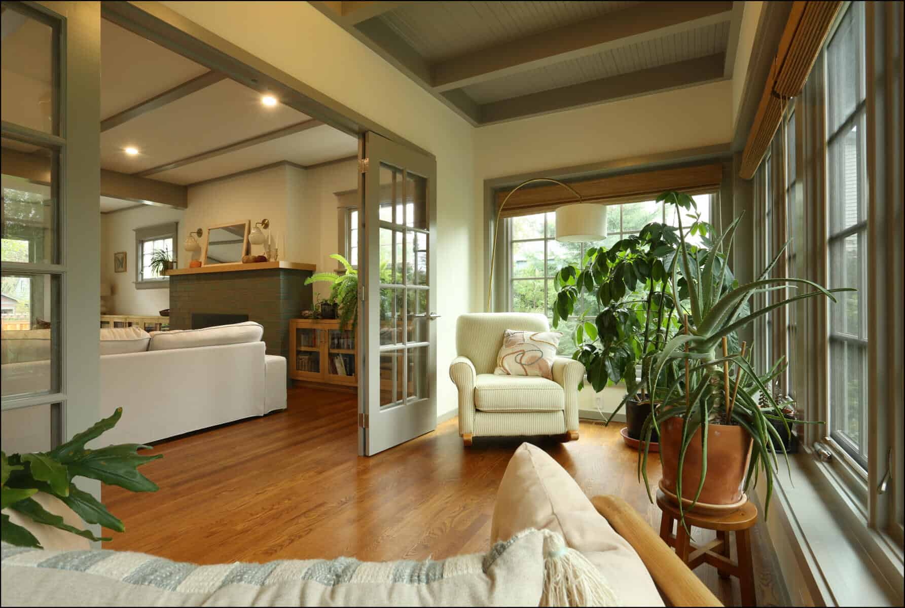 A cozy living room with hardwood floors, large windows, and lush indoor plants. There's a white armchair near a fireplace with a sofa and a wooden coffee table. Cozy living area in a whole house remodel, featuring natural light, hardwood floors, and greenery, showcasing ReCraft Home Remodeling's design expertise in Portland, Oregon.