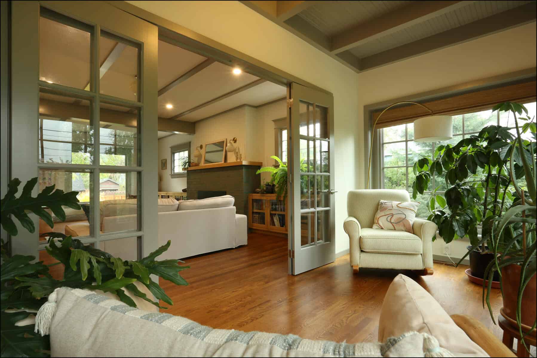 Cozy living room with wooden floors, white sofas, and a beige armchair. Large windows and glass doors bring in natural light. Indoor plants add greenery throughout the space. Cozy living space featuring a bright interior with large windows, a comfortable white armchair, and lush indoor plants, showcasing a whole house remodel by ReCraft Home Remodeling in Portland, Oregon.