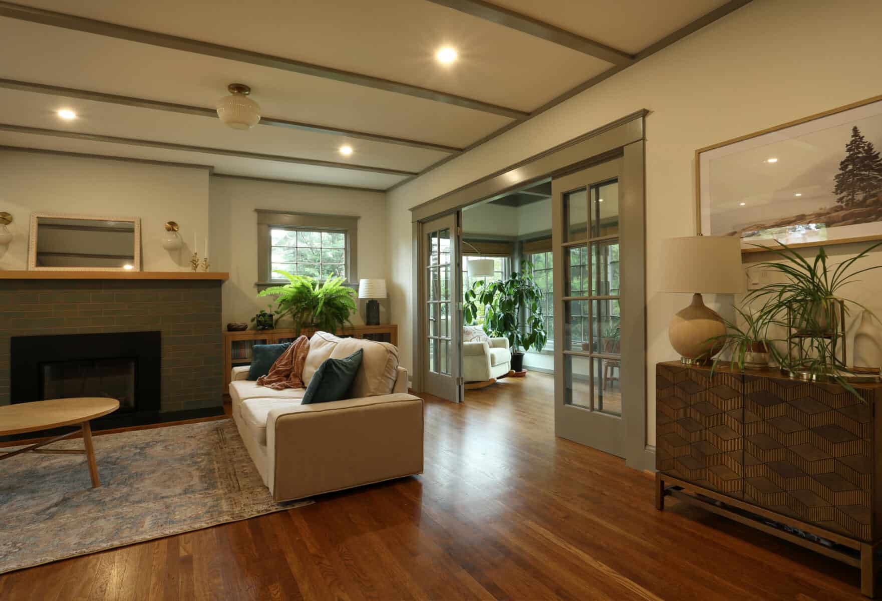 A cozy living room with a sofa, coffee table, and fireplace. Adjacent sunroom with large windows and plants. Hardwood floors and warm lighting complete the space. Modern living room with a cozy sofa, hardwood flooring, and a fireplace, featuring large windows and a glass door leading to a sunlit outdoor space, showcasing ReCraft Home Remodeling's whole house remodel in Portland, Oregon.