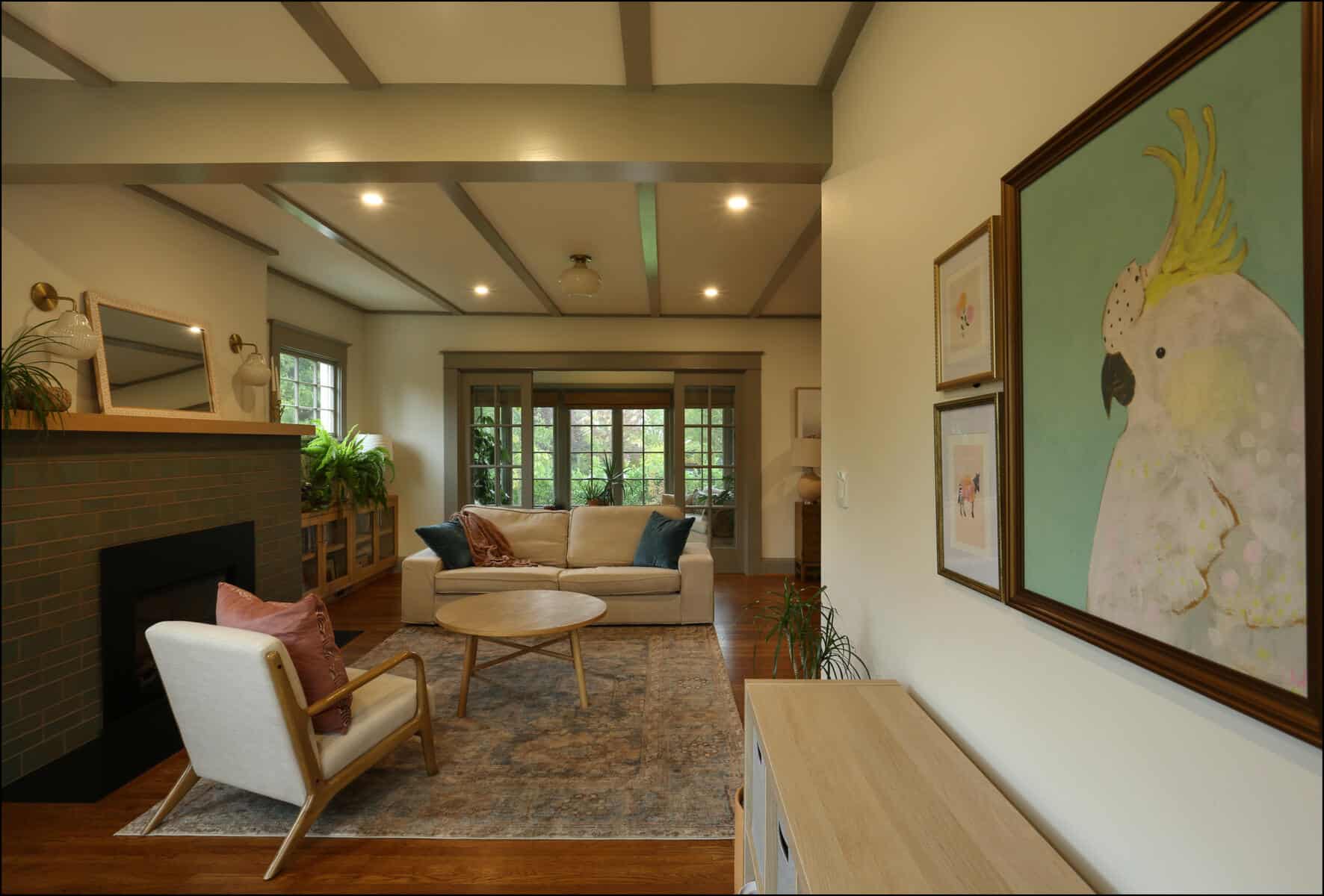 A cozy living room with a sofa, armchair, and round table. A large bird painting hangs on the wall. A fireplace is on the left, and large windows provide natural light. Cozy living room with modern furniture, featuring a white sofa, accent chairs, and a large window, complemented by a colorful bird painting, showcasing ReCraft Home Remodeling's interior design expertise in whole house remodels.
